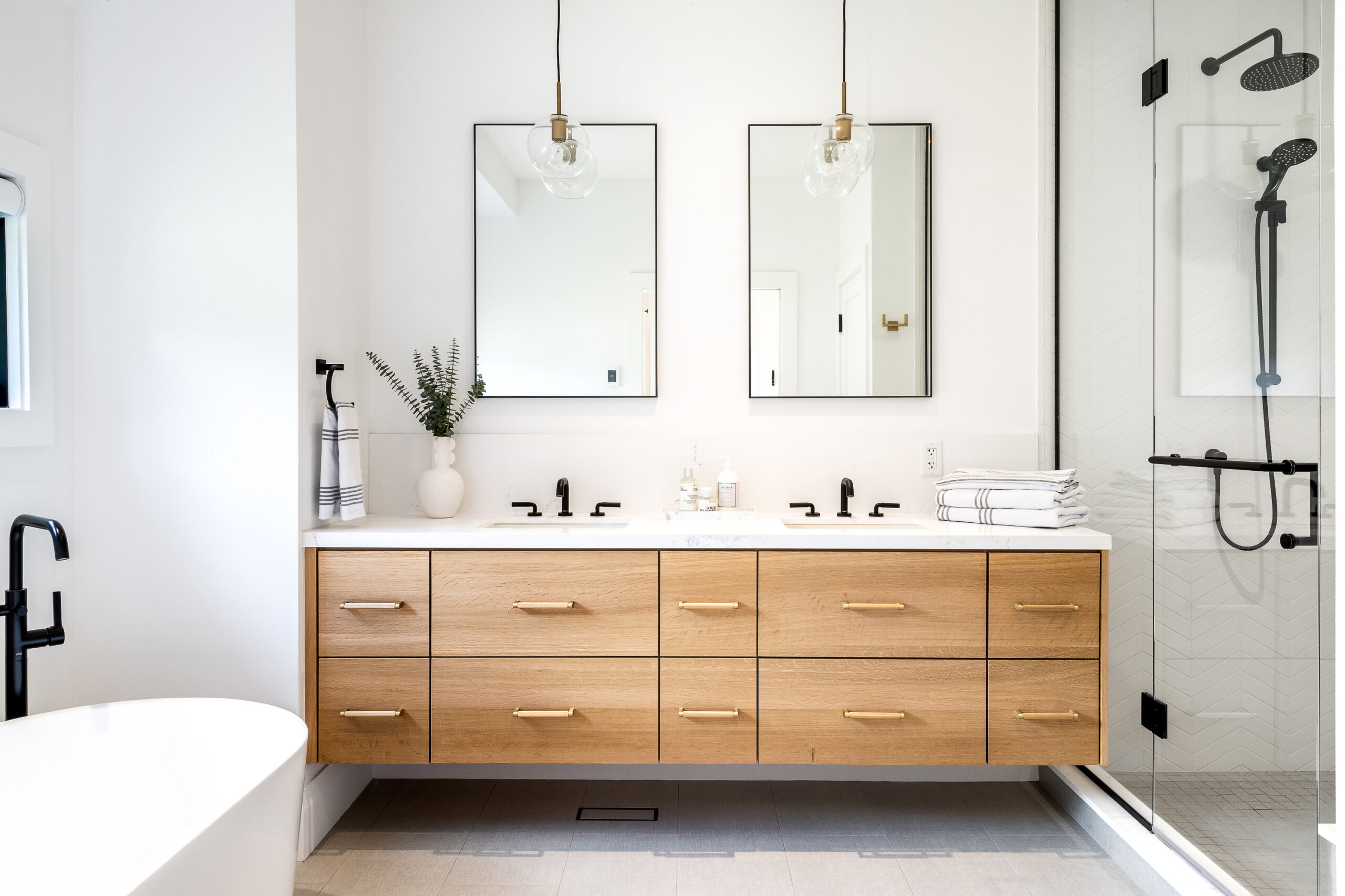 This bathroom features a wood, double vanity with a white countertop, black-finished fixtures, a glass shower and two mirrors hanging over the vanity.